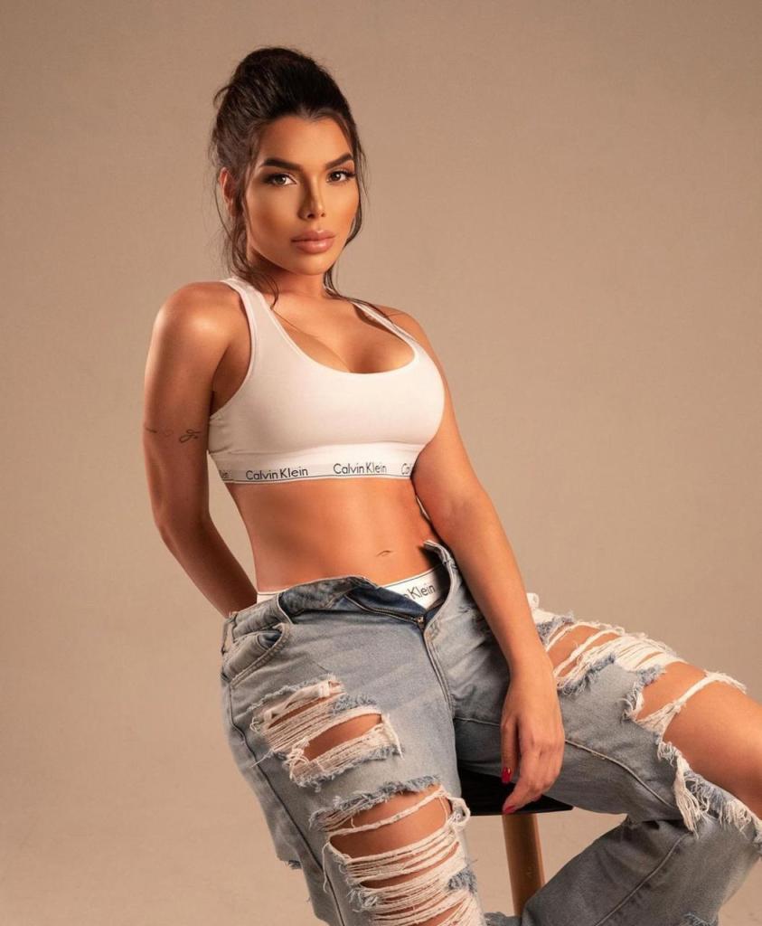 A person wearing a white sports bra and ripped jeans, posing while sitting on a stool against a neutral background.