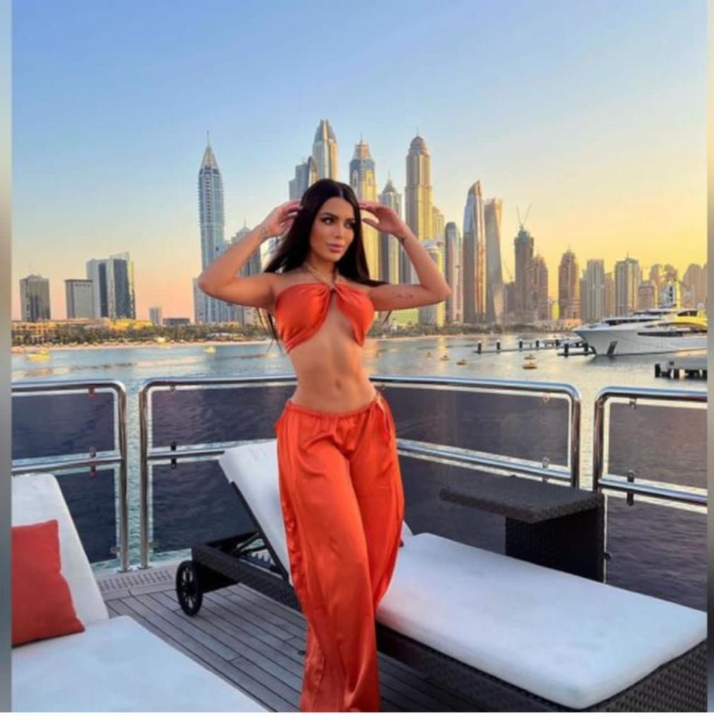 A woman in an orange outfit poses on a yacht with a scenic view of a city skyline during sunset.