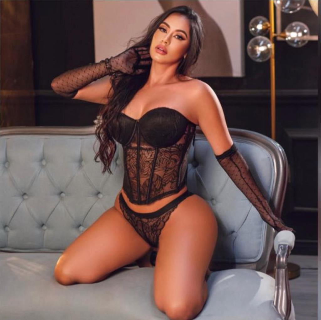 A woman posing on a couch, wearing black lingerie that includes a corset and matching briefs, with long black gloves and styled hair.
