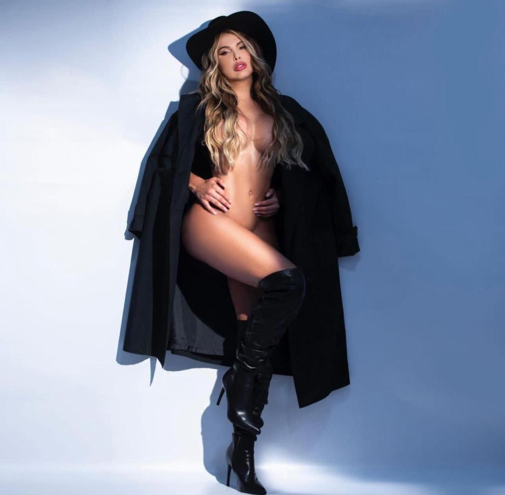 A woman posing confidently in a black coat and knee-high boots against a light blue background.