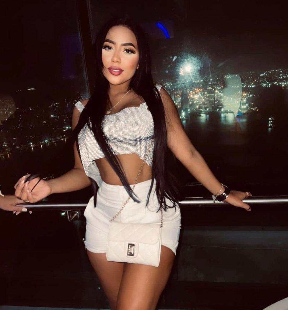 A woman with long black hair poses confidently by a large window at night, wearing a sparkly crop top and white shorts, with a city skyline illuminated in the background.