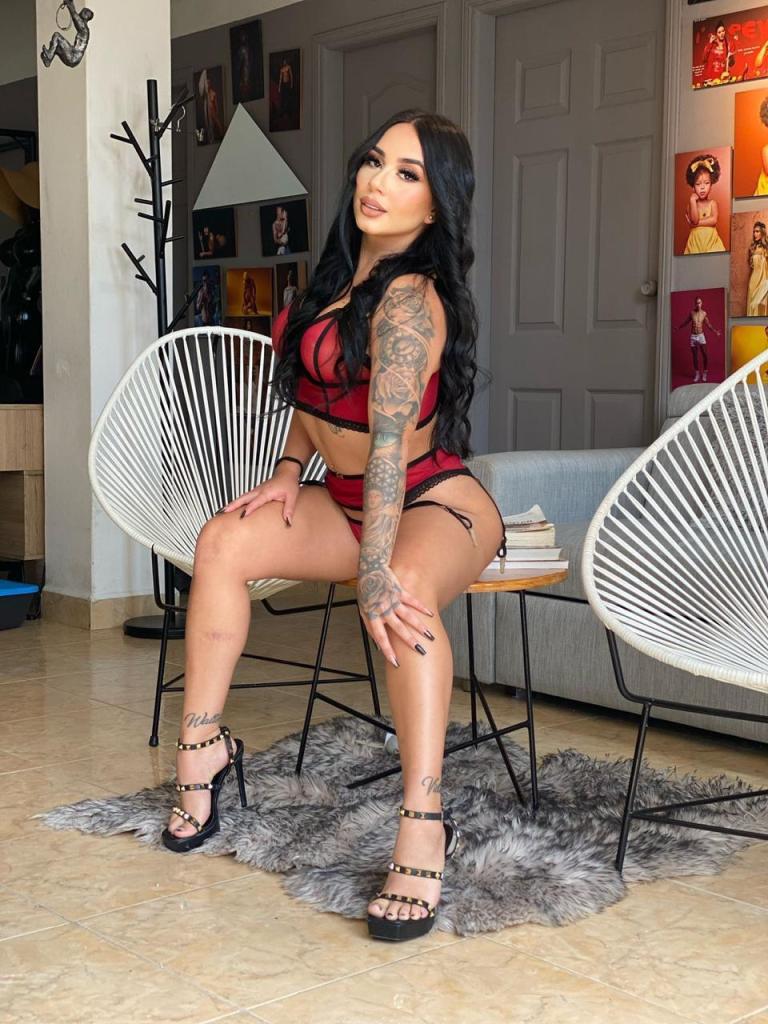 A woman with long dark hair and tattoos poses in a stylish setting, wearing red lingerie and high heels. She is seated on a chair with a fur rug nearby, and artwork is displayed on the walls.